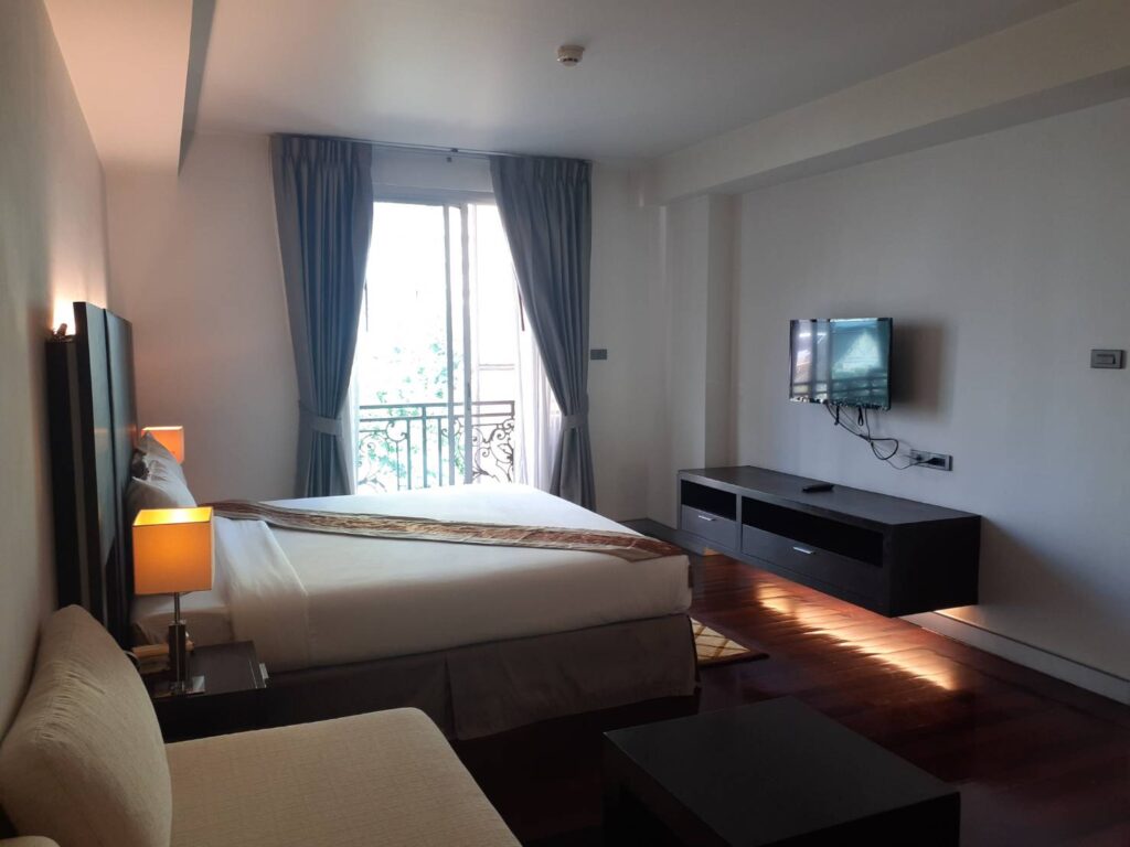 IHバンコクが手配するMona Suiteのホテル客室で、ベッドとテレビ、バルコニーがある室内の様子